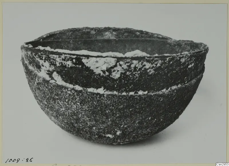 Deep, rounded bowl with a rough, encrusted dark surface and a wide rim, photographed on a pale background.