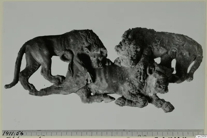 Dark, corroded metal figure group of animals, with a lion on the left, photographed beside a millimetre scale ruler