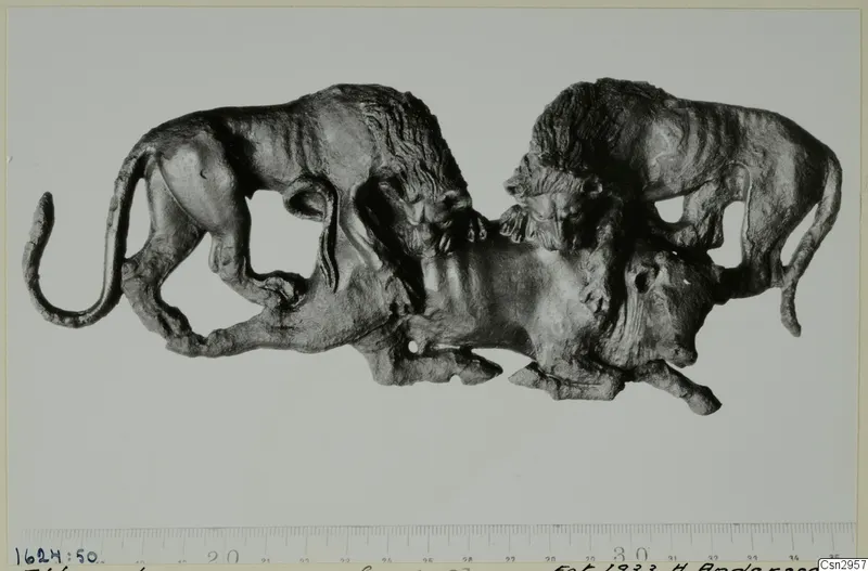Dark metal figure of two maned animals attacking a third animal below, shown with a centimetre scale beneath