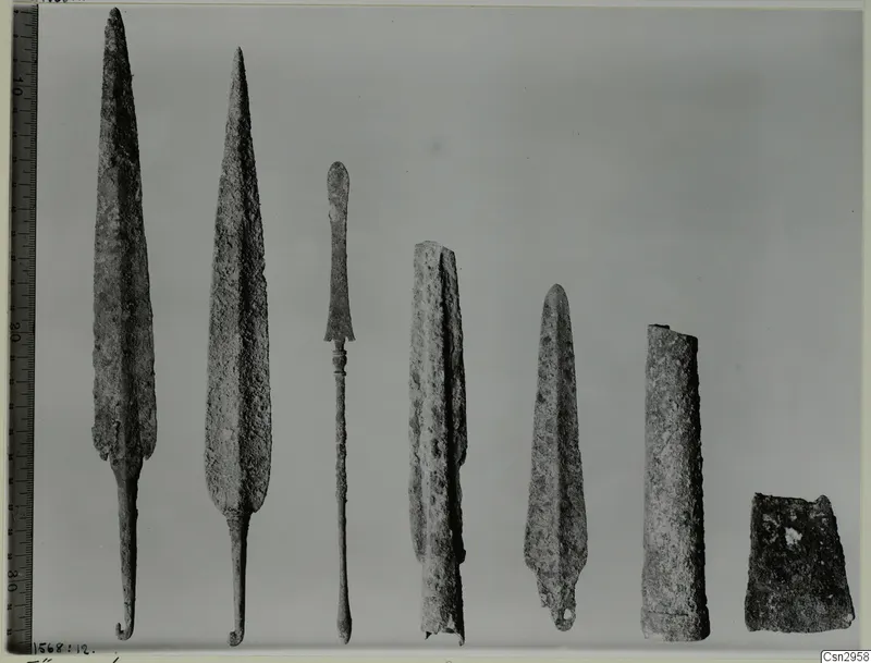 Row of seven corroded metal objects, including two long pointed blades, laid out on a grey background
