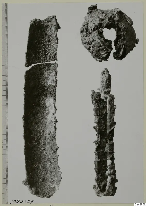Three corroded metal objects on a pale background: a long flat strip broken in two, a ring with a central hole, and a pointed rod.