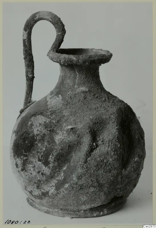 Pottery vessel with a single loop handle rising from its shoulder to the rim, its surface crusted with soil