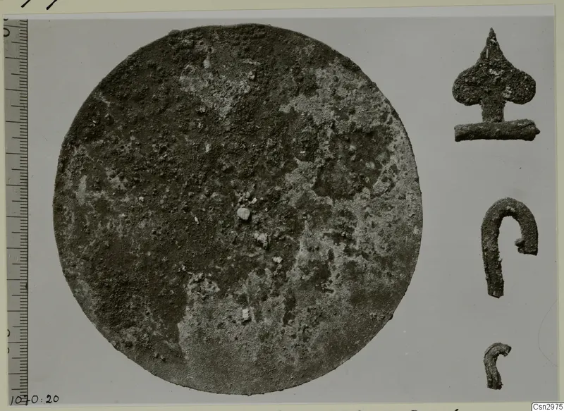 Corroded circular metal disc, with three small corroded metal objects to its right and a measuring scale along the left edge