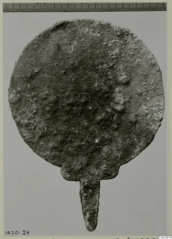 Corroded round metal disc with a narrow tang at its base.