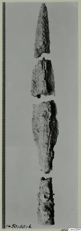 Four corroded metal fragments stacked vertically, the top one tapering to a point, with a measuring scale along the left edge.