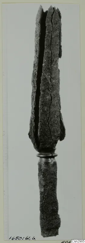 Long corroded metal object with a split, blade-like upper end above a ring collar, shown beside a measuring scale.