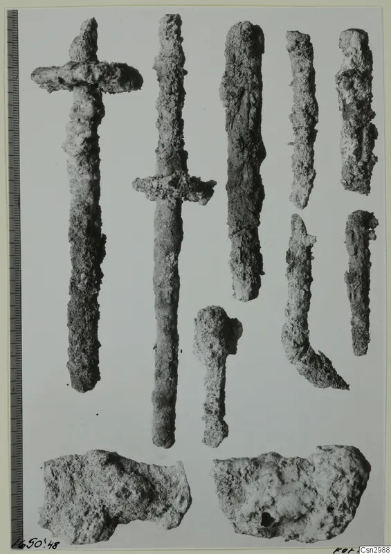 Corroded metal objects before conservation, including two sword-like pieces with cross-guards, laid out beside a scale bar