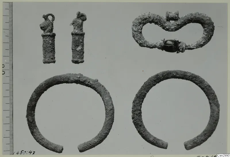 Several small metal objects before conservation: two loop-topped pendants, a figure-of-eight piece with a dark bead, and two open bangles.