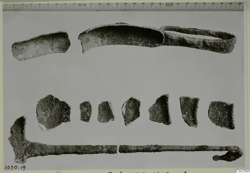 Corroded metal fragments laid out in rows beside a ruler, including a curved strip and a long rod with a hooked end.