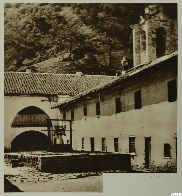 Whitewashed building with a bell hung in a wooden frame at Machairas, Troodos, with a wooded hillside behind.