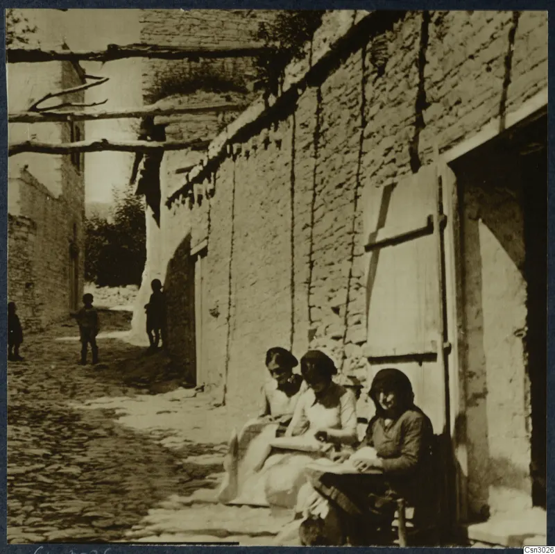 Cobbled village lane with three seated women against a stone wall and children standing in the street