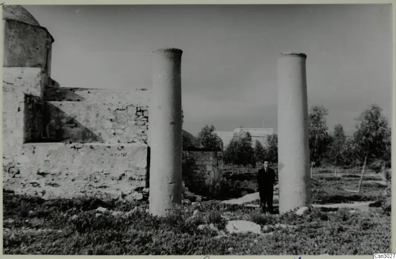 Man in a dark suit standing between two stone columns beside a ruined building, Paphos