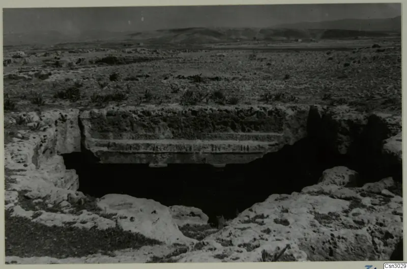 Dark rock-cut opening under an overhanging rock ledge in open scrubby ground, with low hills behind