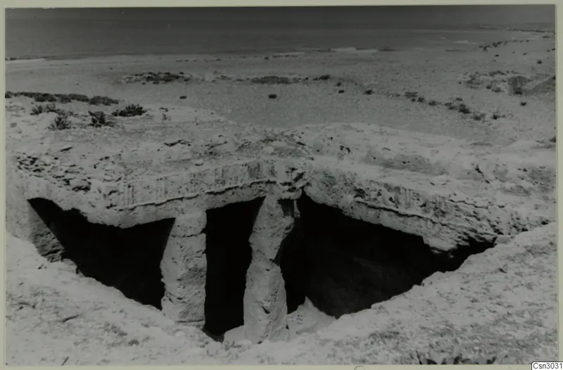 Flat-roofed rock structure resting on two pillars, with dark openings beneath it, on sandy ground above a calm sea.