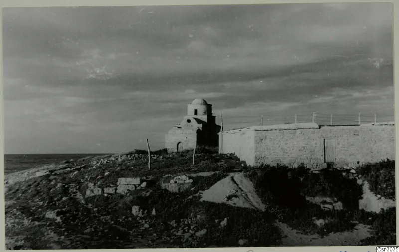 Small domed building beside a long whitewashed wall and wire fence on a rocky coastal rise