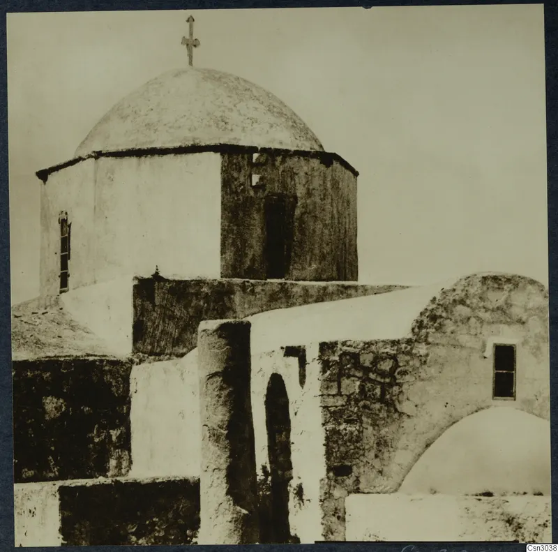 Whitewashed domed church with a cross on top and a small window, with a stone wall and arched doorway in front.