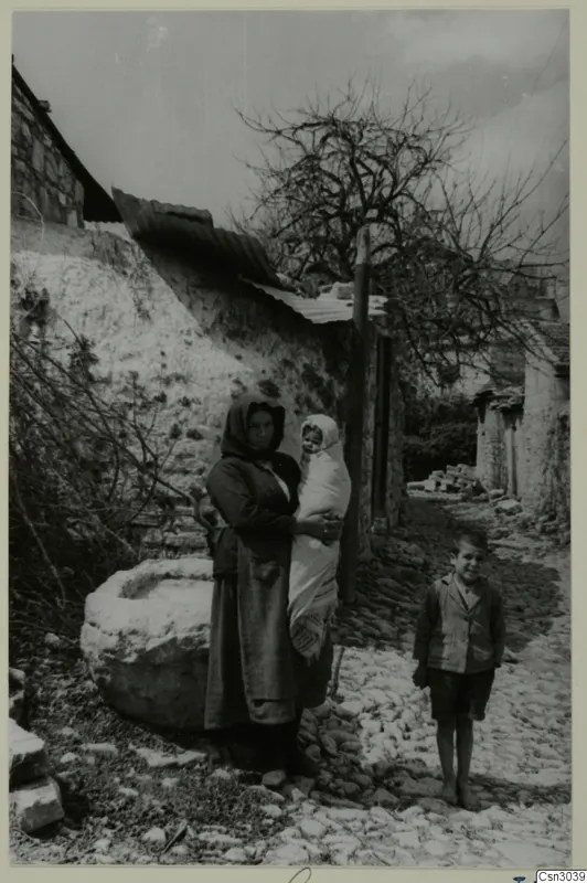 Woman holding a baby and a boy standing on a cobbled lane beside a large stone basin and a stone wall, Paphos