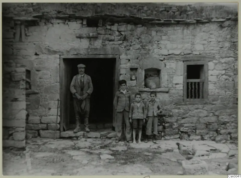 Man and three boys standing at the doorway of a stone building, with a bird on the paved ground in front