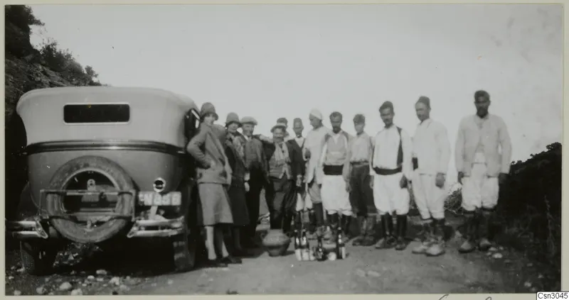 Group of people standing beside an old car on a dirt road, with a large pot and bottles near their feet