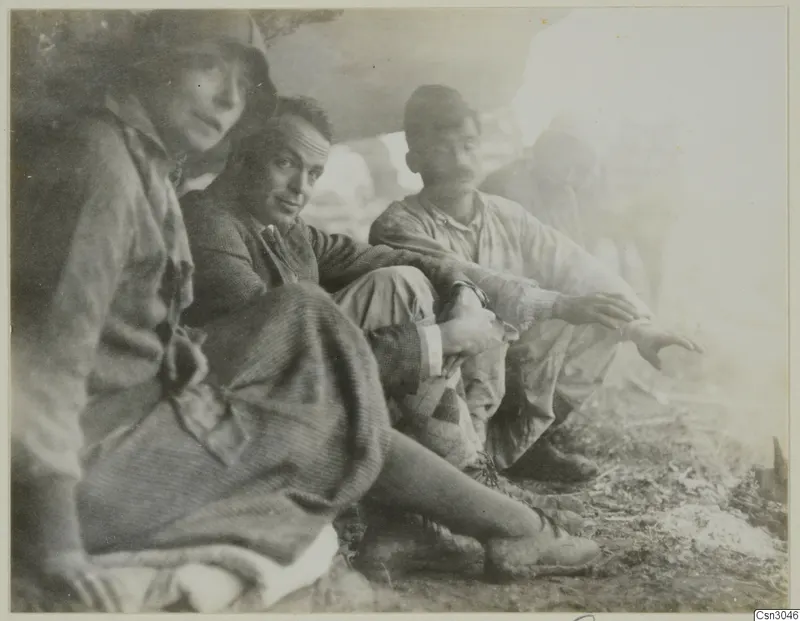 Four people sitting and resting on dry ground at a site, one in a hat and one in a long skirt, in a bright hazy setting.