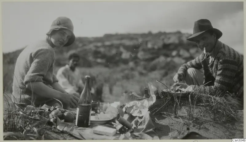 Picnic on a grassy hillside: a woman and a man in hats with a glass bottle, food and paper wrappings in the foreground