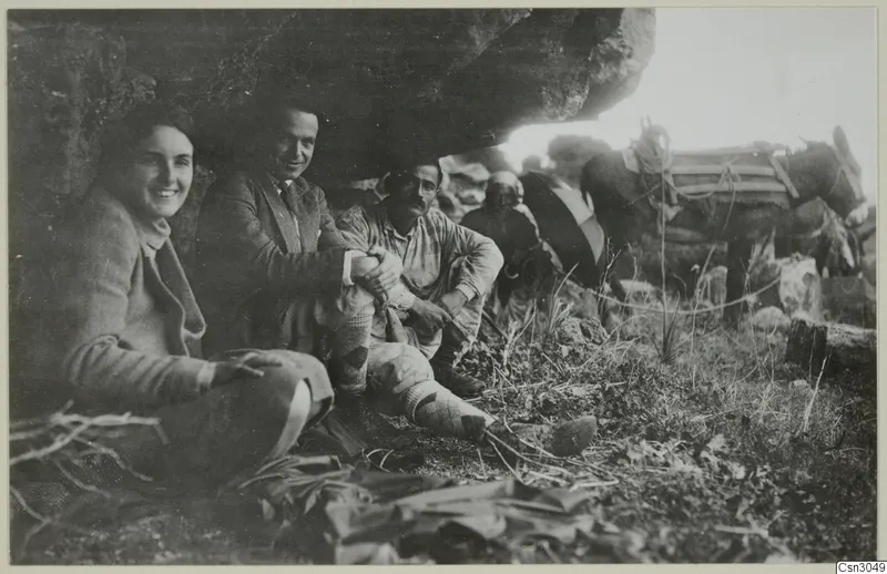 People and a loaded donkey resting under a large rock overhang, with grass and low plants in the foreground.