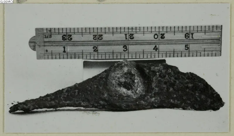 Corroded metal object with a rounded boss and tapering point, lying beside a folding ruler for scale