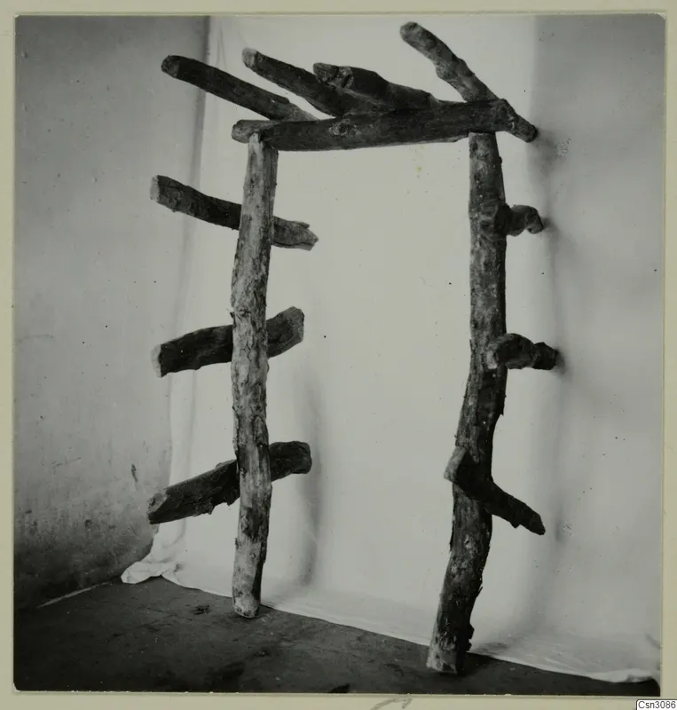 Wooden frame of rough, bark-covered branches standing on a floor against a white cloth, from the Cypriote Mining group.