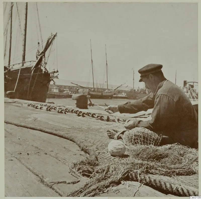 Man in a cap mending a fishing net on a harbour quay, with masted sailing boats moored behind him