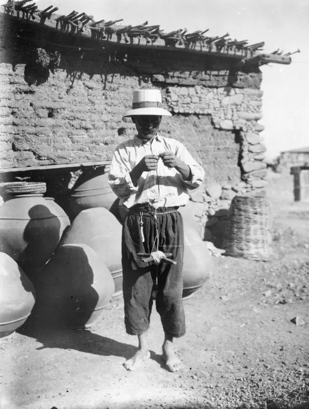 Rope-maker twisting net cord beside large clay jars and a mud-brick wall at farmstead A, Klirou