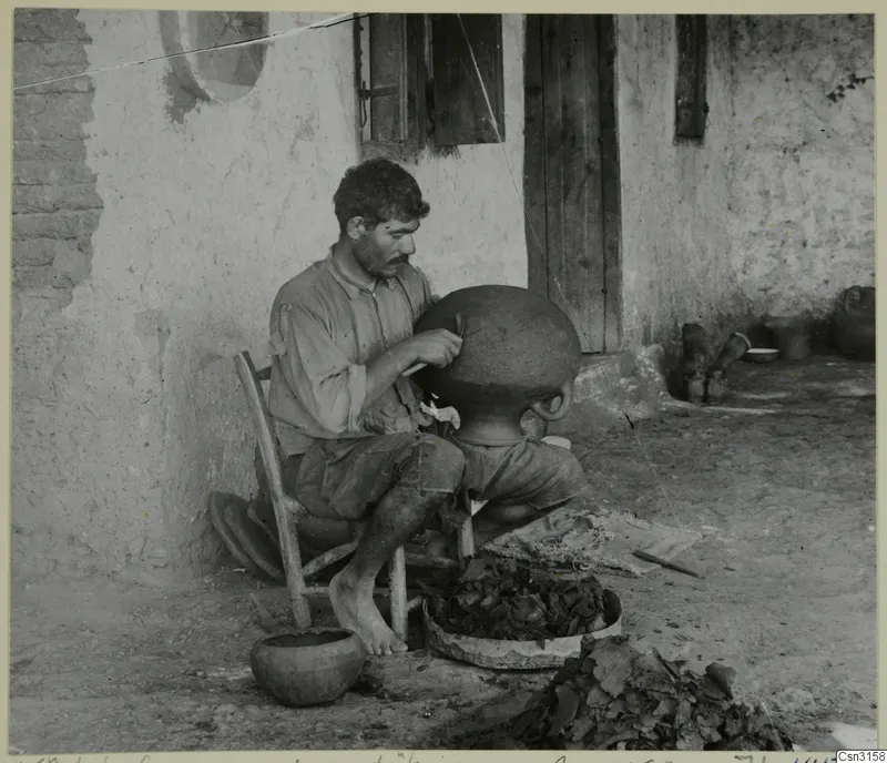 Man shaping a large clay vessel with a wooden knife, seated outside a whitewashed wall with a wooden door