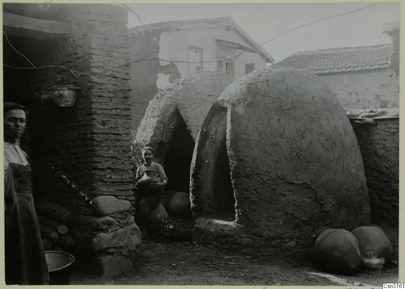 Domed pottery kilns with arched openings in a yard, with a man holding a jar and a woman at the left edge.