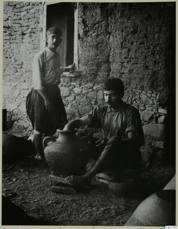 Potter turning a large clay jar outside a stone and mud-brick building, with a second man standing in the doorway behind him.
