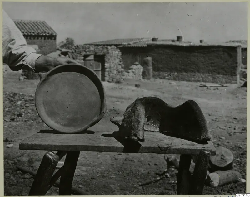Round fired clay dish and a curved clay windbreak on a wooden bench, with village houses behind at Kliron