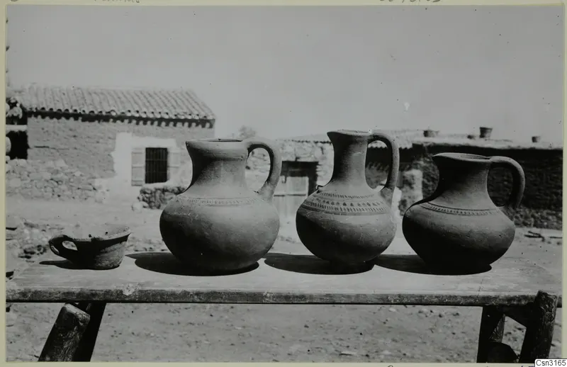 Three jugs and a small handled cup on a rough wooden table, with village houses behind, Kliron