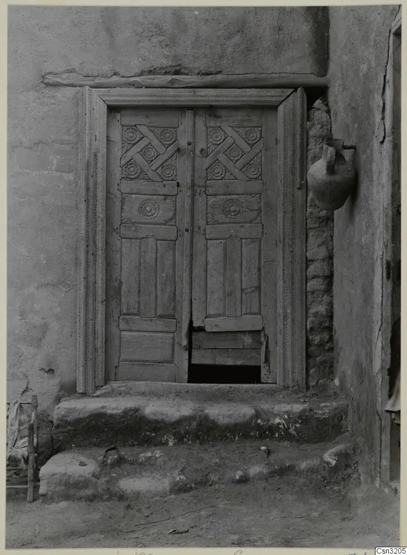 Carved wooden double door in a plastered wall, with worn stone steps below and a clay pot hanging to the right