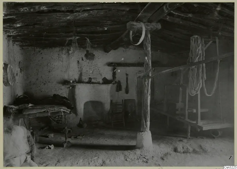 Interior of a plastered room with a log ceiling, a central wooden post, a low frame with bedding and a wooden frame on the right.