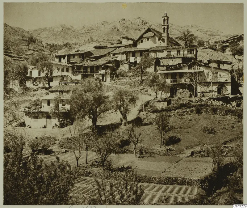 Mountain village of plastered houses with tiled roofs and a tall bell tower, above terraced fields