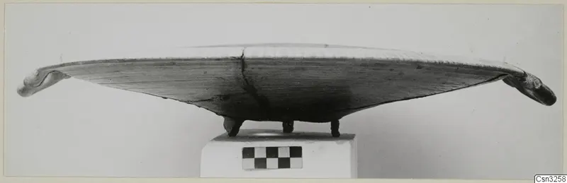Shallow pottery vessel with upturned knobbed ends, standing on three small feet, photographed side-on on a plinth.
