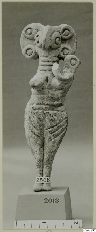 Standing terracotta figurine with a wide head decorated with round discs and one arm raised, shown on a block base