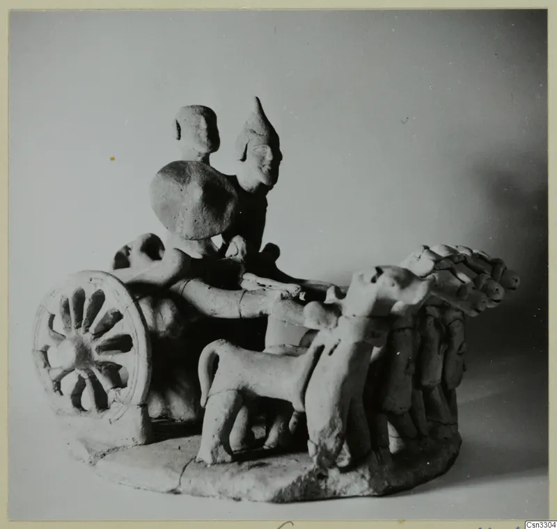 Small terracotta model of two standing figures on a wheeled frame, with a spoked wheel at left and animals in front.