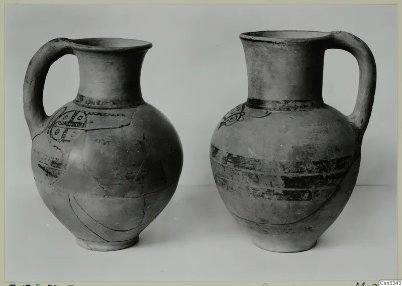 Two handled pottery jugs with dark painted bands and motifs, photographed side by side against a grey background.
