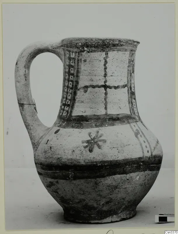 Painted pottery jug with a strap handle, dark bands and panels of dots and cross lines on a pale body.