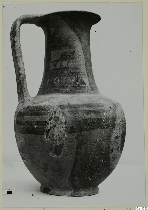 Painted pottery jug with a strap handle and dark banded decoration, photographed as a find against a grey backdrop.
