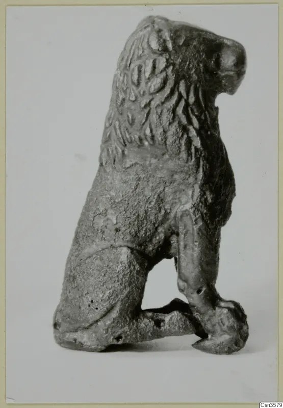 Small bronze figure of a seated animal with a textured mane, shown in side view on a pale backdrop