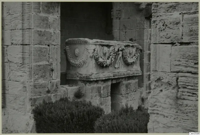 Stone sarcophagus with a carved garland band, standing in a recess between stone block walls