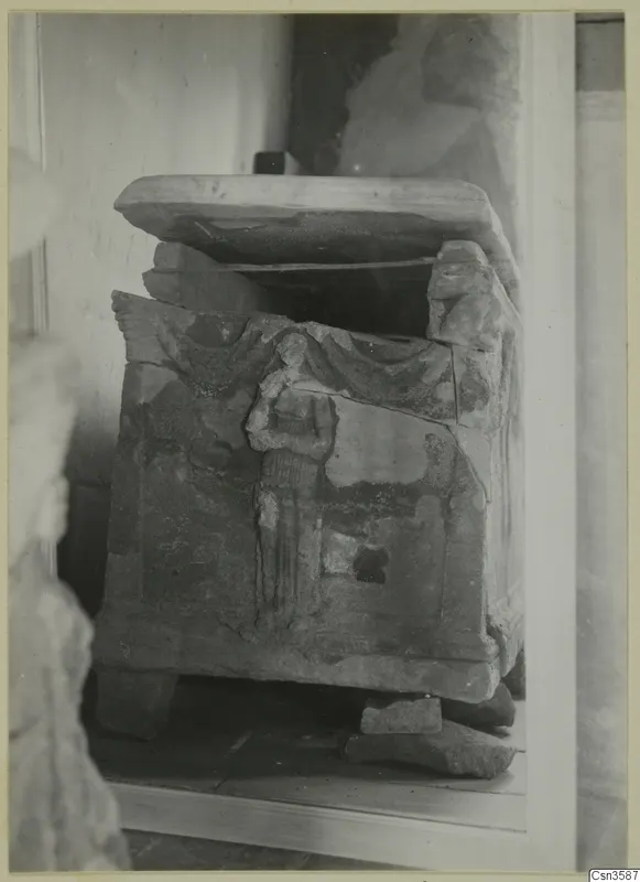 Stone sarcophagus with a carved standing figure on its front, lid slid askew, photographed indoors against a plain wall