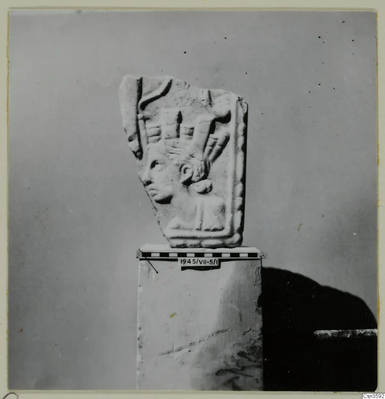 Relief fragment showing a head in profile with an elaborate headdress, set on a stone block with a scale bar beside it.