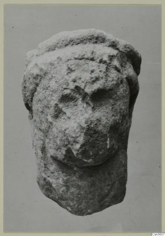 Limestone statuette fragment of a head with a band across the top, face worn away, shown against a grey backdrop