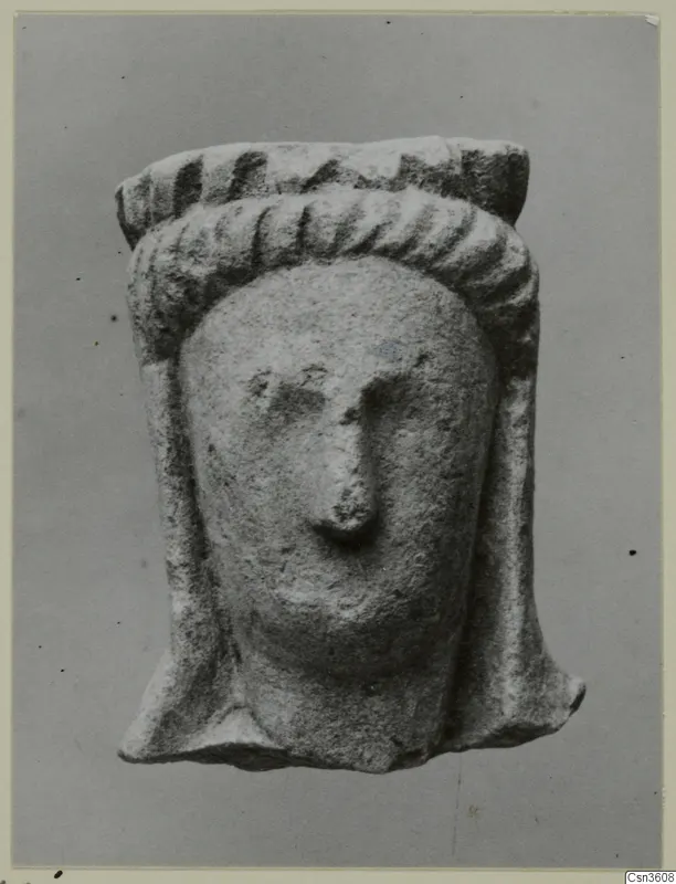 Limestone figurine of a head with ridged headdress and long side locks, shown against a plain grey backdrop.